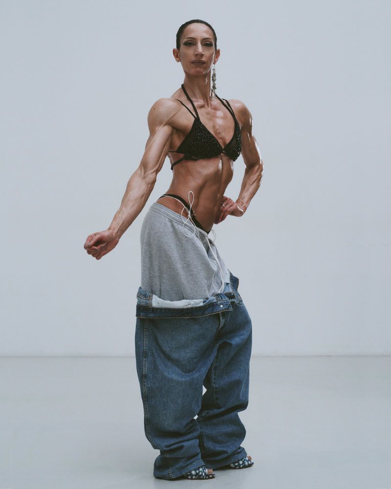 Muscular adult female in a bikini top and oversized jeans posing in a photo studio with a neutral background and diffused lighting.