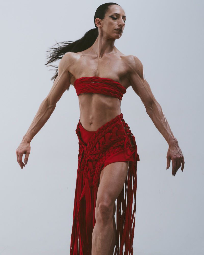 Adult female posing in a woven red top and skirt with fringe, in a studio, with arms outstretched and hair pulled back in a ponytail.