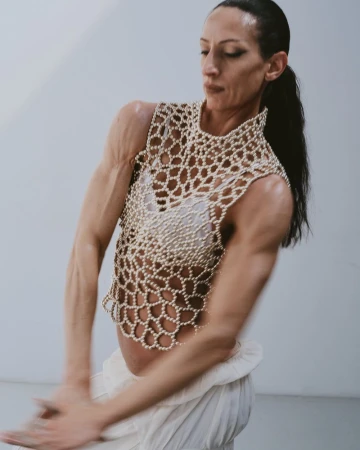 Adult woman poses in motion, wearing a pearl-beaded top and white skirt against a plain studio backdrop, captured in soft, natural light.