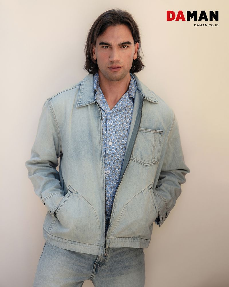 Portrait of a male model in a light denim jacket and patterned shirt against a neutral wall, showcasing fashion and casual style.
