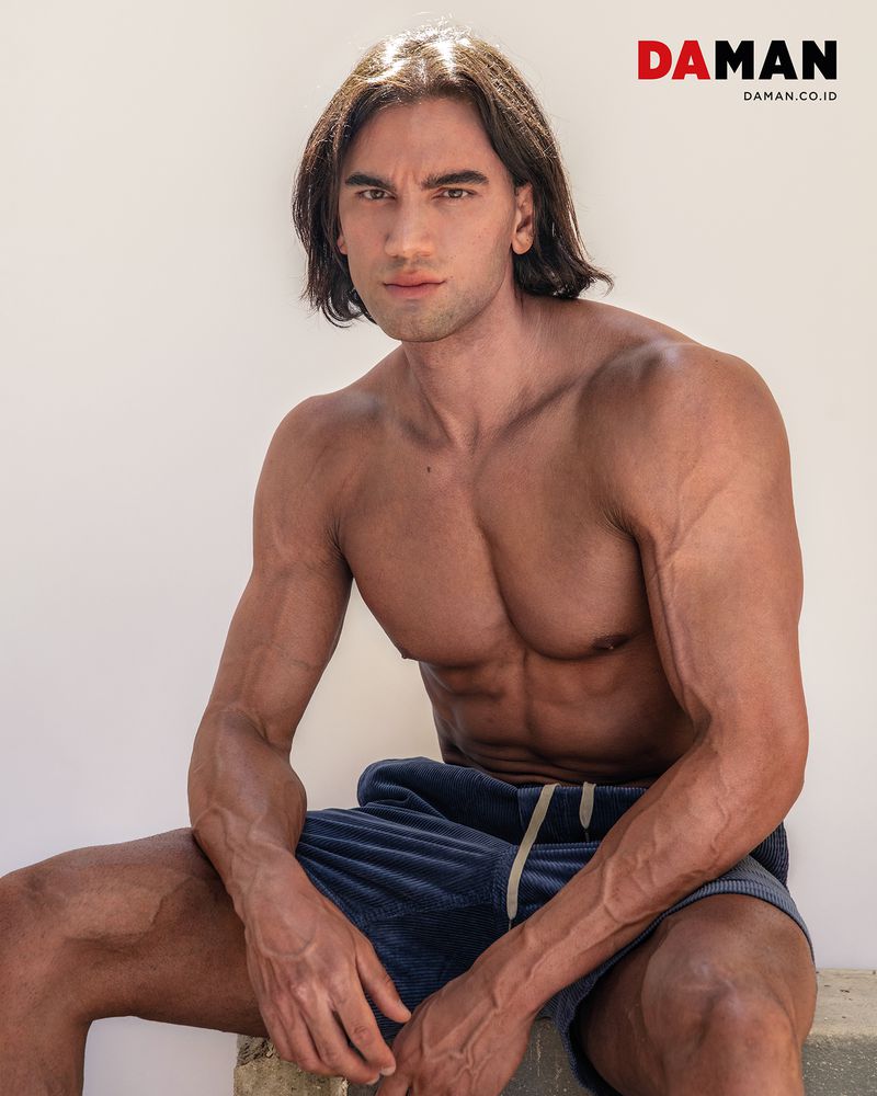 Model Chand Smith sitting shirtless in navy drawstring shorts against a plain off-white studio background.
