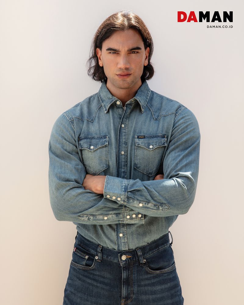 Portrait of a dark-haired man wearing a denim shirt and jeans, standing indoors with his arms crossed against a neutral wall.