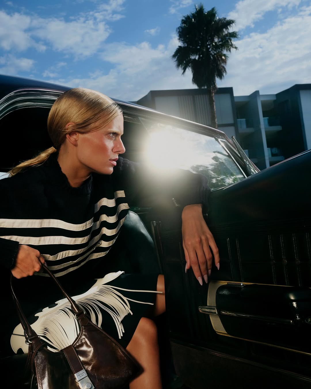 Blonde woman wearing striped sweater leaning out of vintage car window holding leather handbag