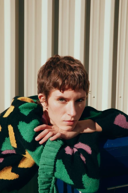 Woman with short hair wearing a patterned green, yellow, and pink sweater with her hands under her chin and a metal background.