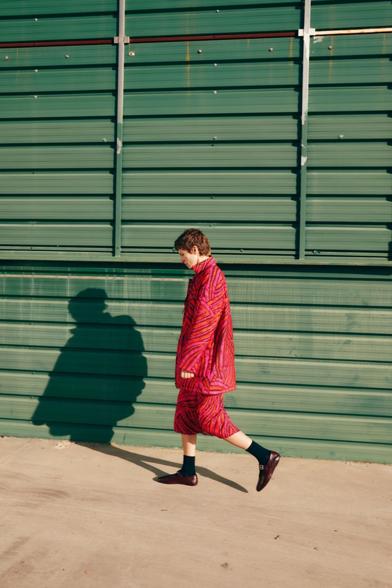 A young adult strides confidently against a corrugated green wall, wearing a patterned red and pink jacket and skirt with brown shoes.