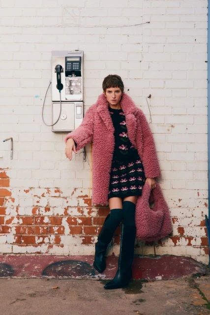 A woman in a pink coat, black dress, and boots poses against a brick wall with a payphone, showcasing a fashion-editorial aesthetic.