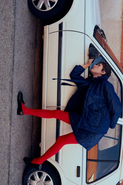 Woman leans against white car wearing red tights and a blue quilted jacket in an urban environment with brick and concrete.