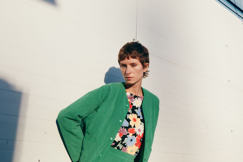 Adult woman wearing a green button-down jacket and floral top poses in front of a white textured wall in a medium shot.