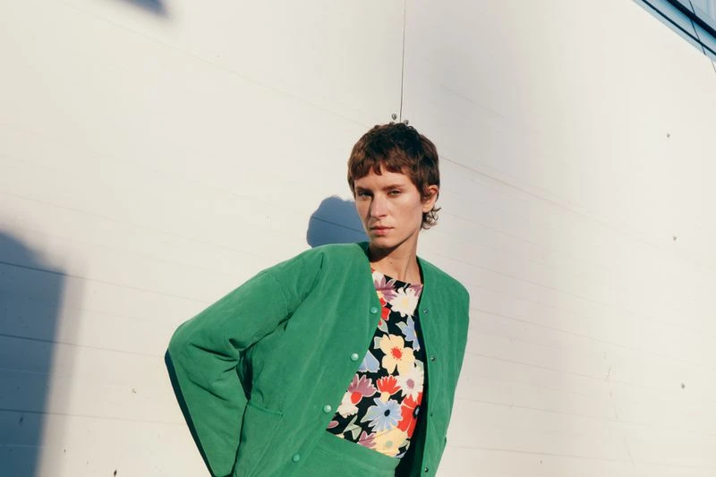 Adult woman wearing a green button-down jacket and floral top poses in front of a white textured wall in a medium shot.