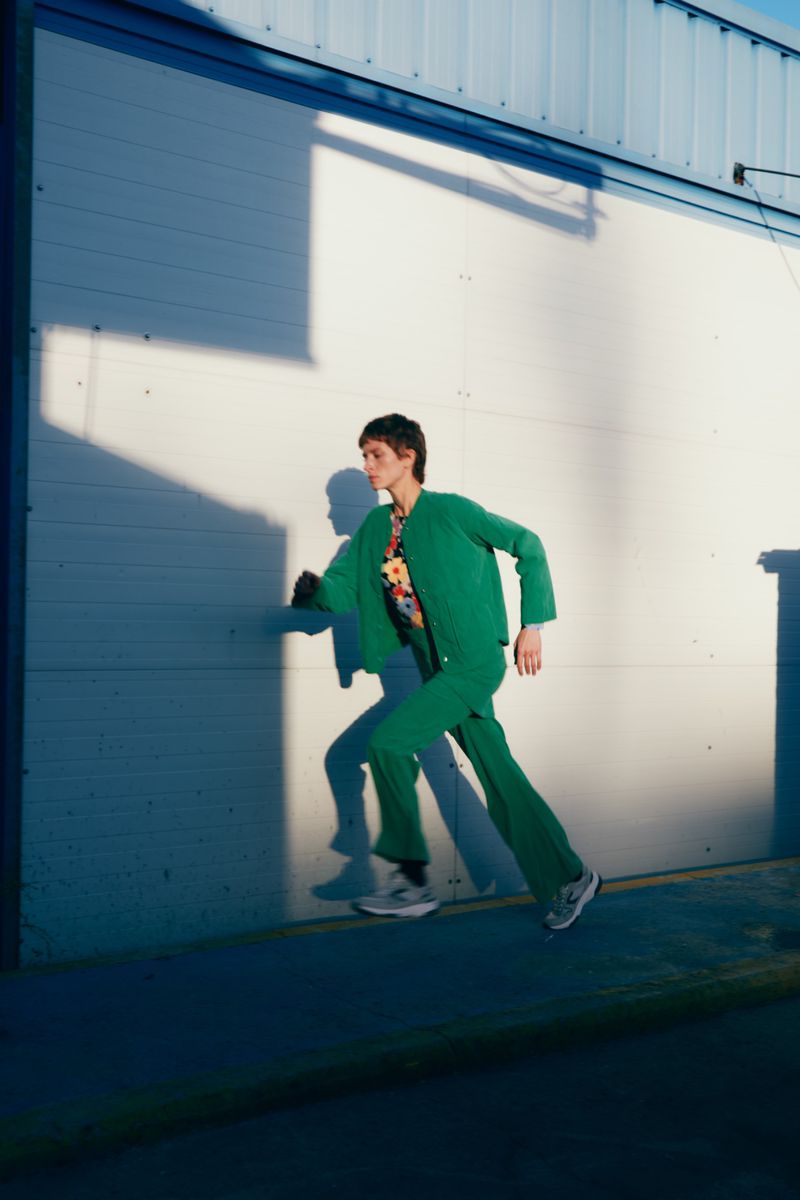 Person in green suit runs against white wall with shadows. Sunny urban scene shows full-body shot of running adult near building.