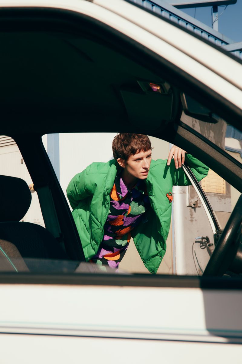 An adult leaning out the open window of an old white car in an outdoor urban setting, wearing a bright green puffer jacket.