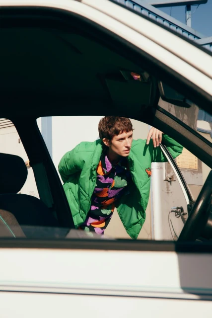 An adult leaning out the open window of an old white car in an outdoor urban setting, wearing a bright green puffer jacket.