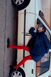 Model wearing a navy quilted jacket and bright red tights posing leaning against a vintage white car on an asphalt street.