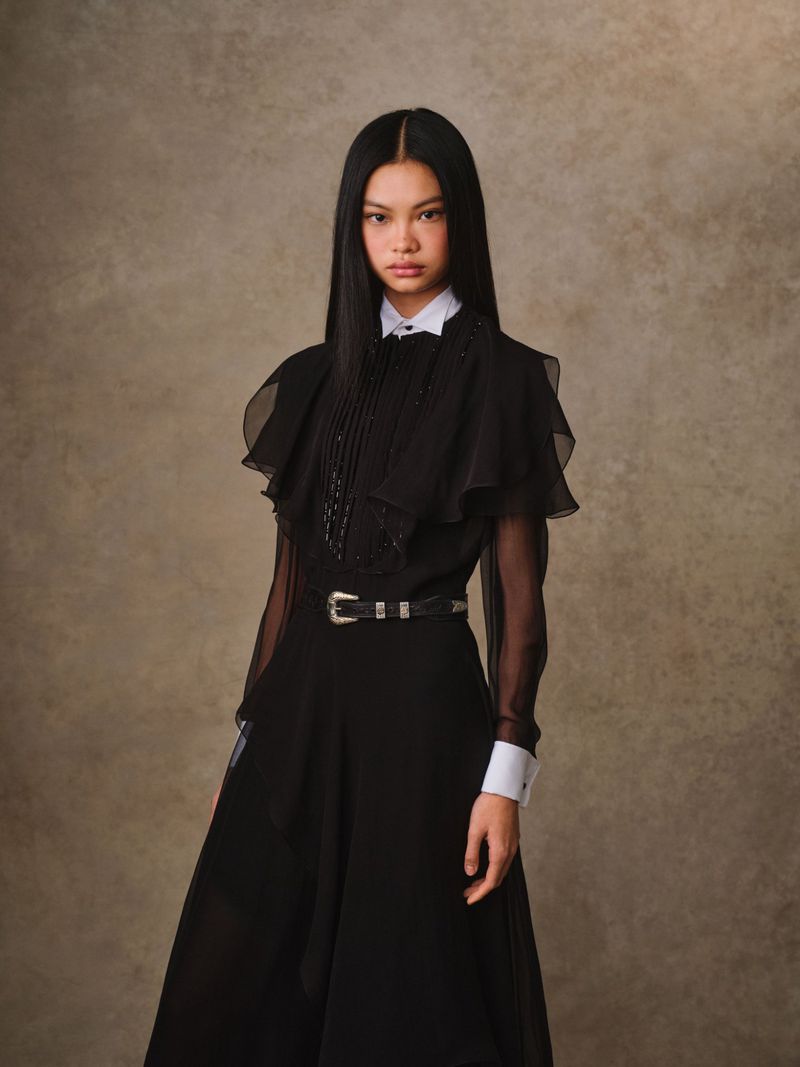 A fashionable young woman in a black dress with sheer sleeves, white collar, and matching belt stands against a mottled brown backdrop.