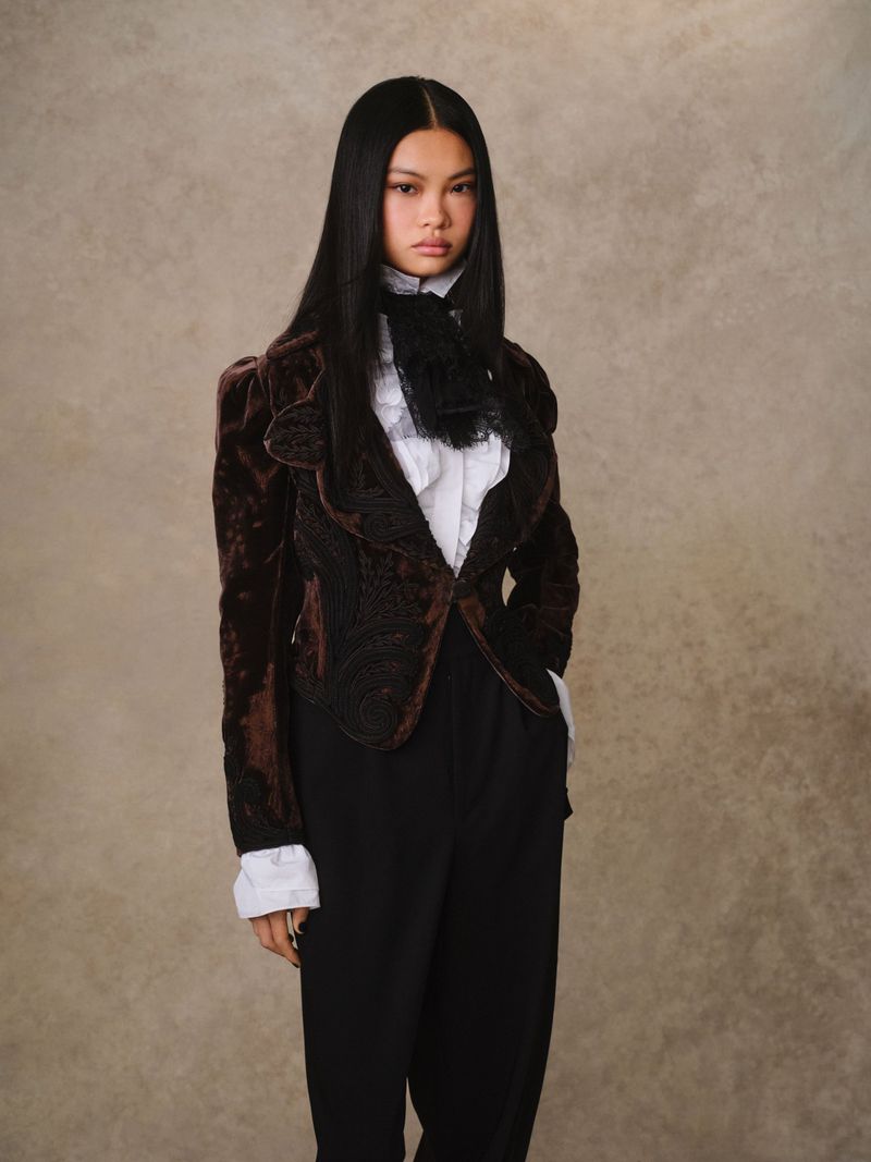 Portrait of an Asian adult female wearing a brown velvet jacket, white blouse, and black pants against a neutral backdrop in studio lighting.