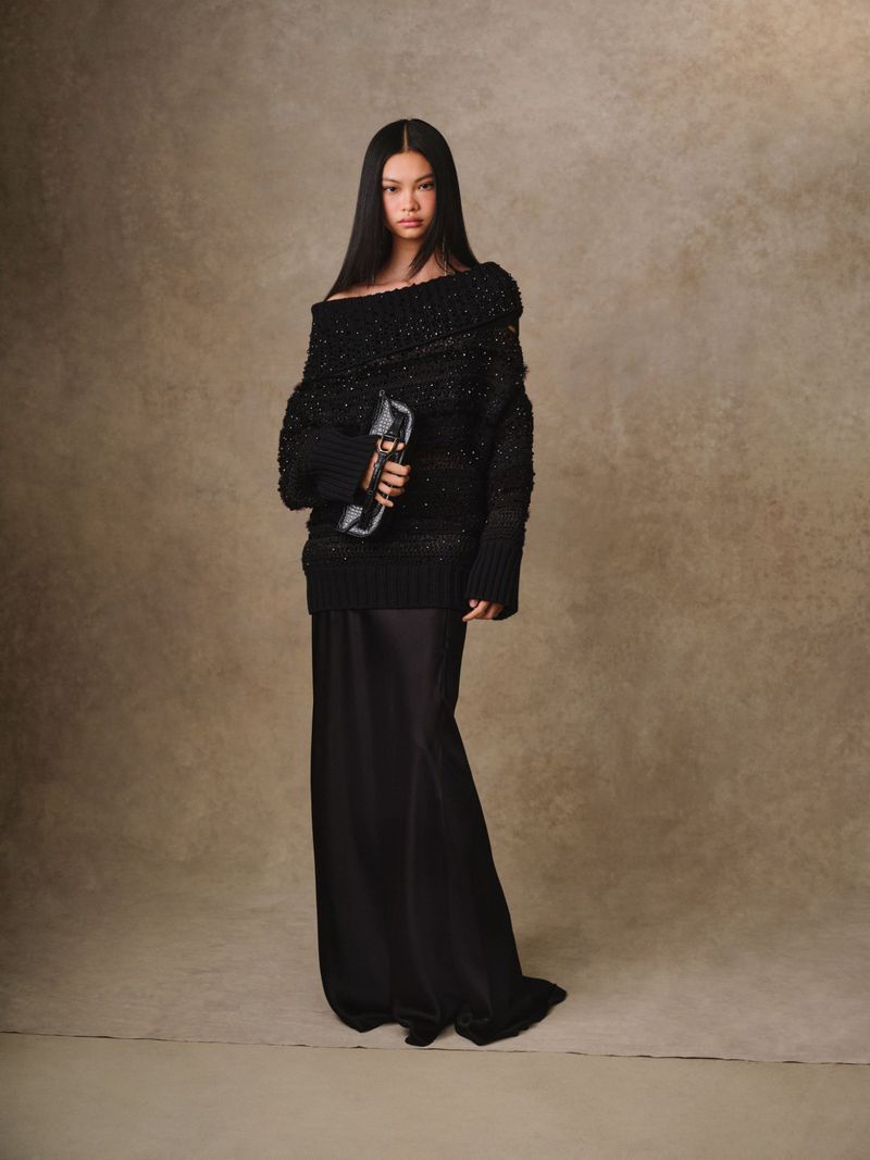 Brunette woman poses in an off-the-shoulder black sweater and long black skirt, holding a clutch in a studio with a neutral backdrop.