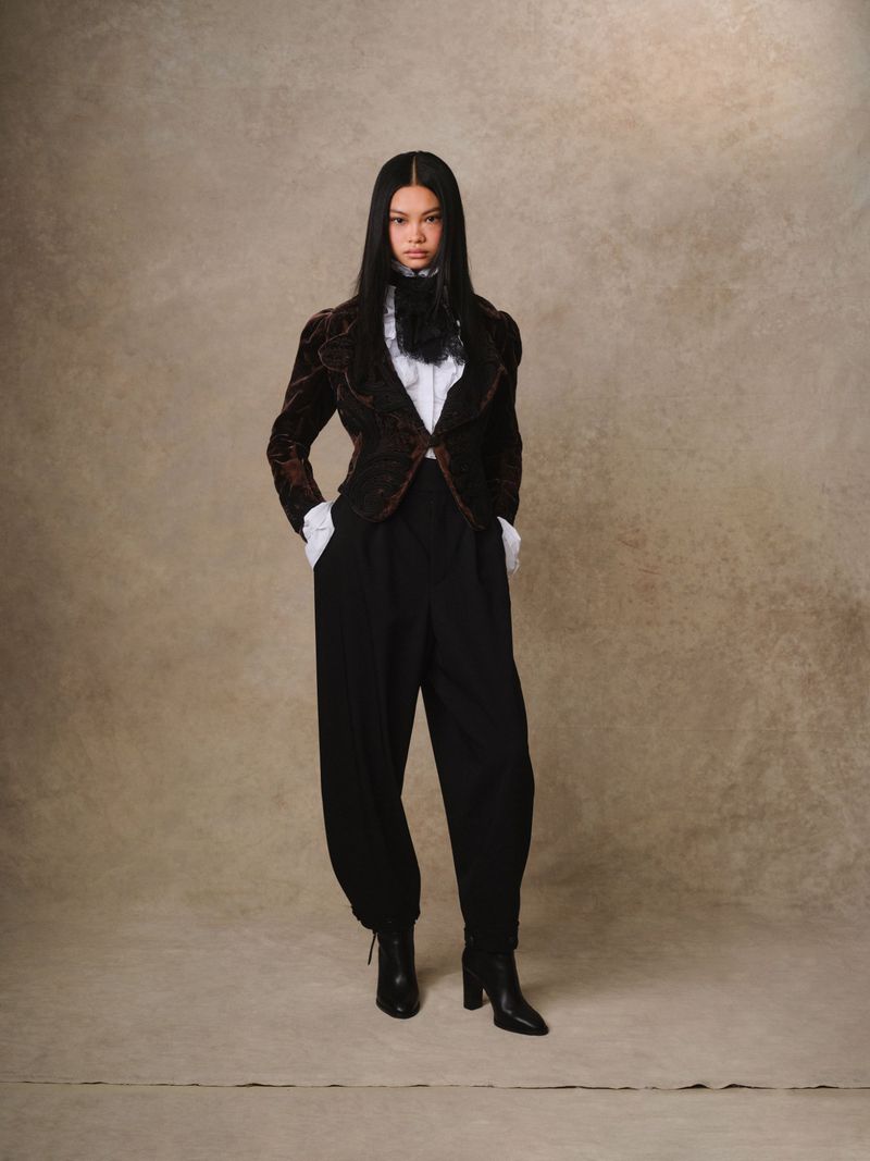 A woman with long, dark hair is posing in a brown velvet jacket, black trousers, and black boots against a neutral background.