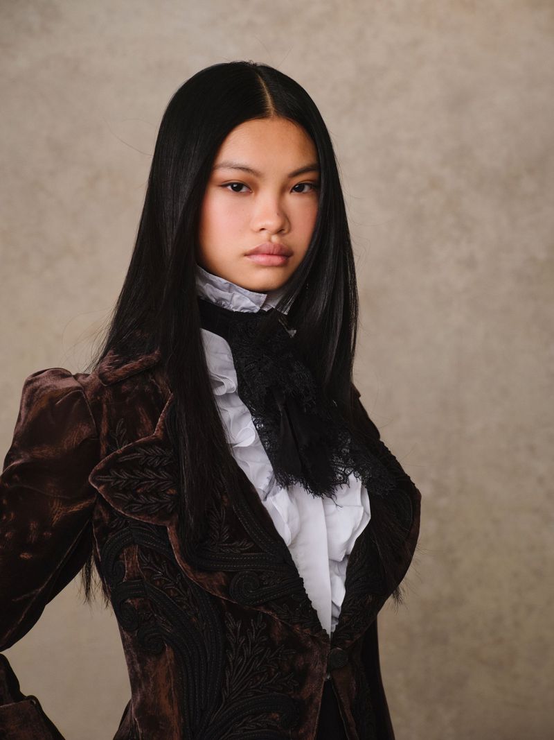 Close-up portrait of a woman with dark hair wearing a vintage-inspired brown velvet blazer and a ruffled white top against a neutral backdrop.