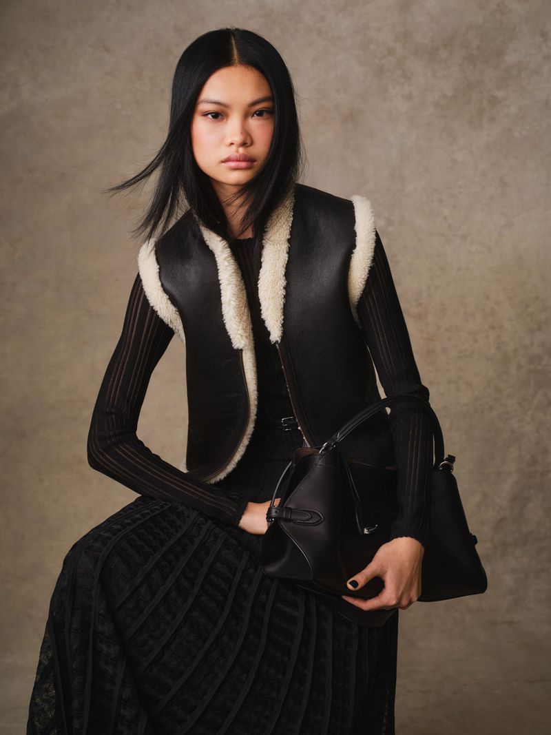 Portrait of a stylish Asian woman wearing a black vest and holding a black leather bag against a mottled beige background.
