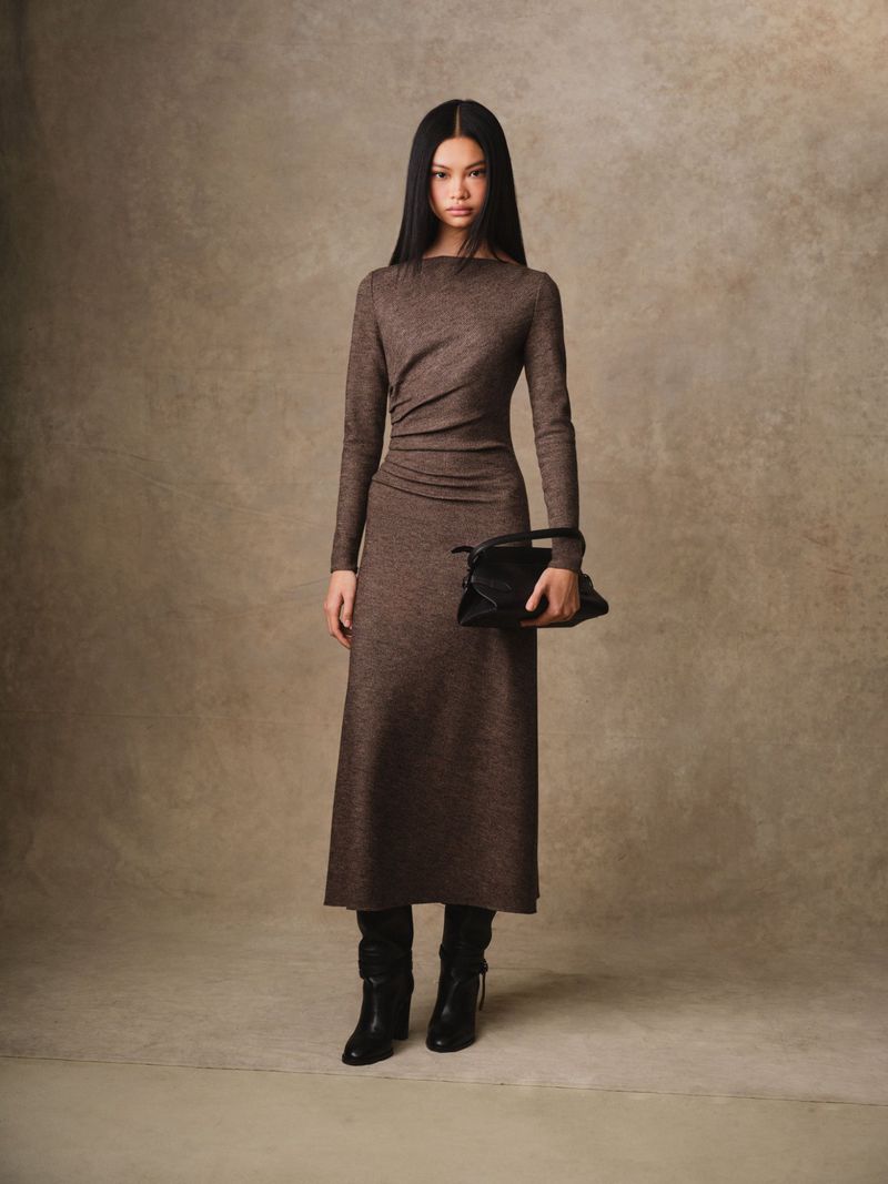 A full-length shot of a fashionable adult female wearing a long-sleeve brown dress, holding a black handbag, and posing in a studio.