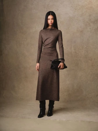 A full-length shot of a fashionable adult female wearing a long-sleeve brown dress, holding a black handbag, and posing in a studio.