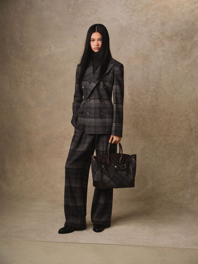 Fashion editorial shot of a woman in a grey plaid suit with a black turtleneck, holding a brown leather handbag against a neutral backdrop.