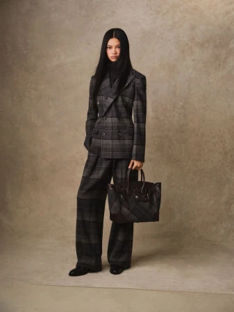 Fashion editorial shot of a woman in a grey plaid suit with a black turtleneck, holding a brown leather handbag against a neutral backdrop.