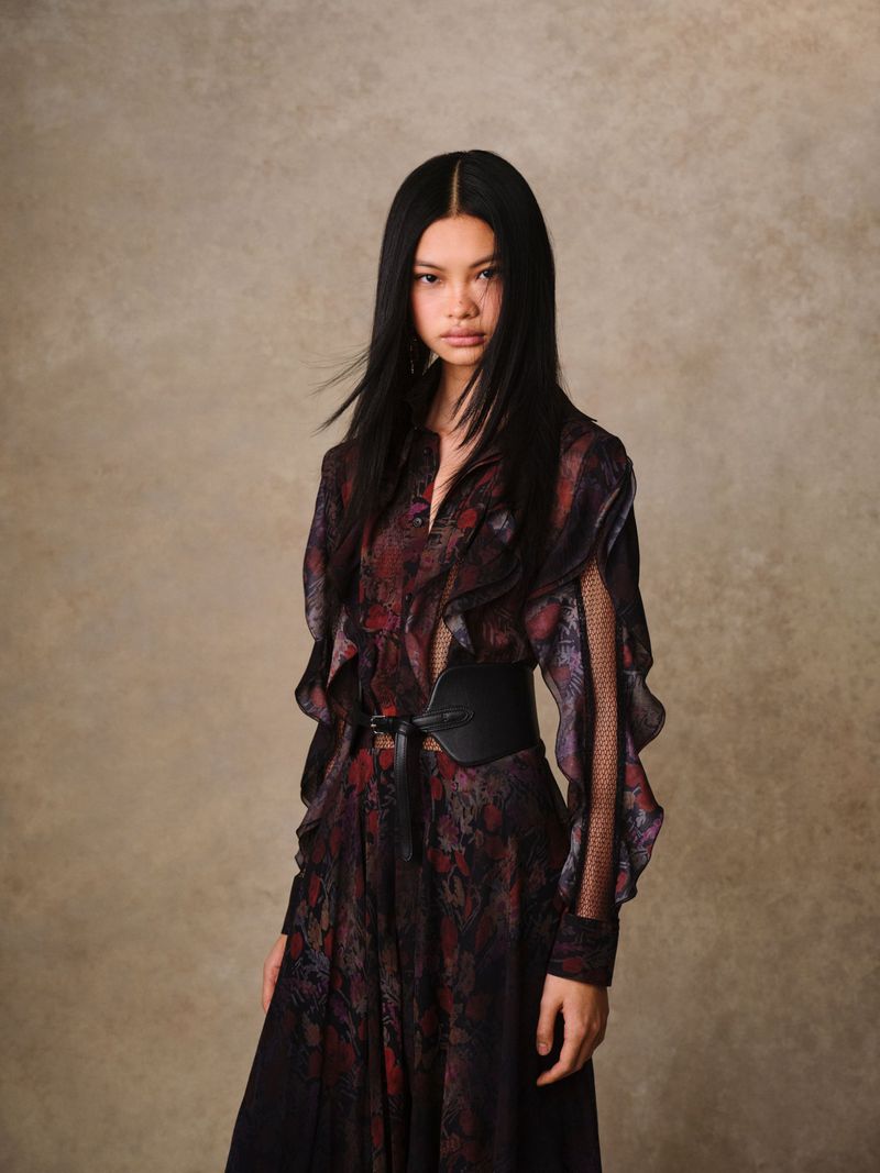 Portrait of an Asian woman with long black hair, wearing a floral dress and a black waist belt, set against a neutral backdrop.