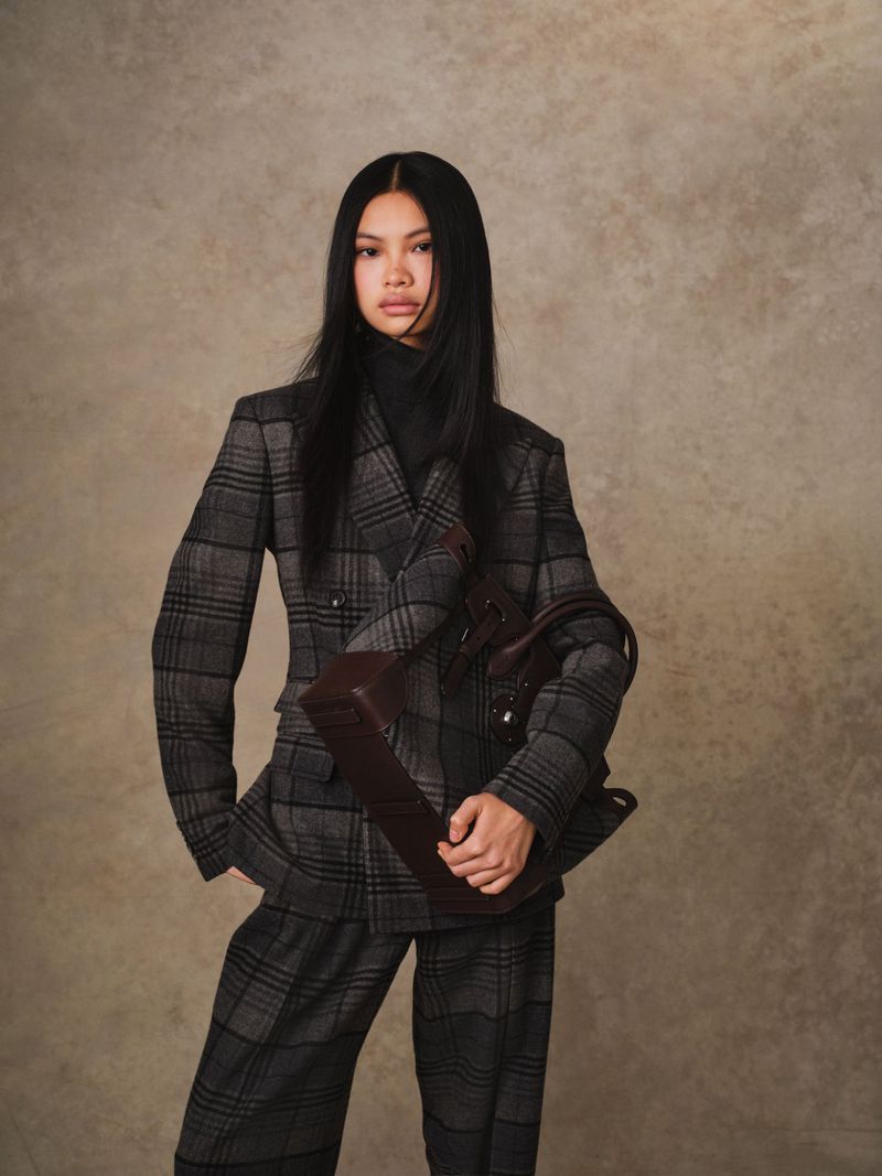 Woman in a gray plaid blazer and matching pants, holding a burgundy handbag, poses against a mottled brown background in a studio setting.