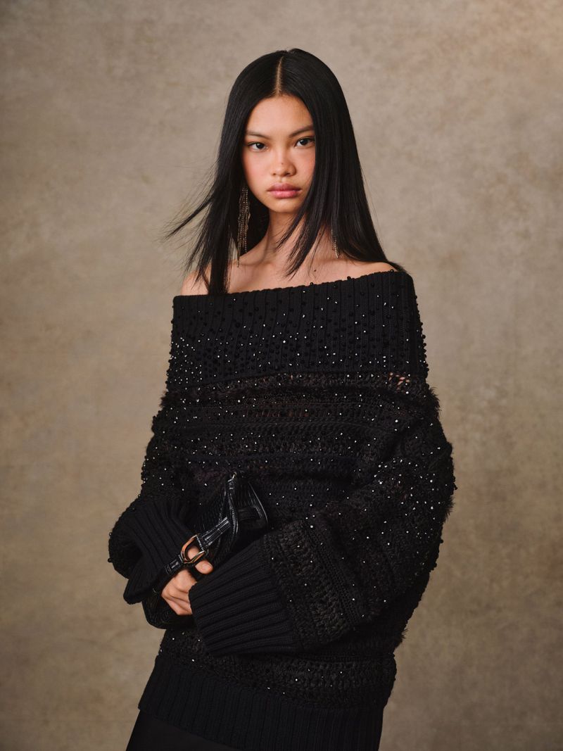 Fashion portrait of a stylish Asian woman in a black knit off-the-shoulder sweater, posing in front of a neutral background.