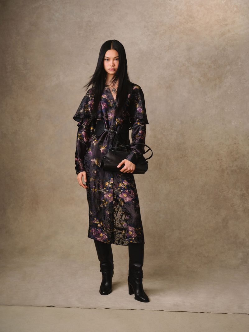 Full-length shot of a slender woman in a floral dress, black boots, and handbag against a mottled backdrop in a fashion editorial style.