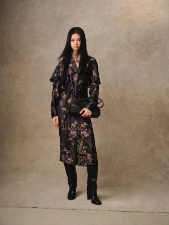 Full-length shot of a slender woman in a floral dress, black boots, and handbag against a mottled backdrop in a fashion editorial style.