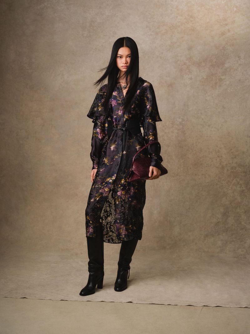 A full portrait of an Asian woman in a patterned trench coat with black leather boots, holding a burgundy clutch in a beige studio.