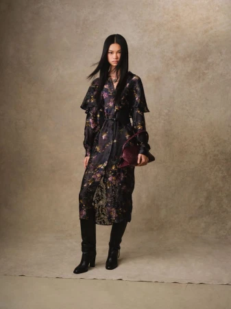 A full portrait of an Asian woman in a patterned trench coat with black leather boots, holding a burgundy clutch in a beige studio.