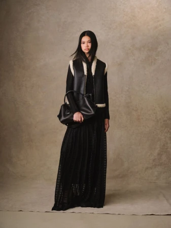 Model Yue Gu in a black shearling-lined vest and long pleated black skirt holding a leather handbag against a textured backdrop.