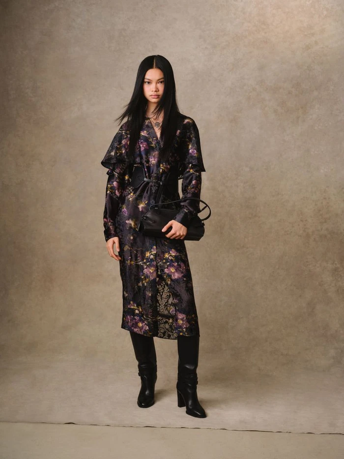 Model Yue Gu poses in a dark floral midi dress, knee-high black boots, and a black handbag against a mottled beige backdrop.
