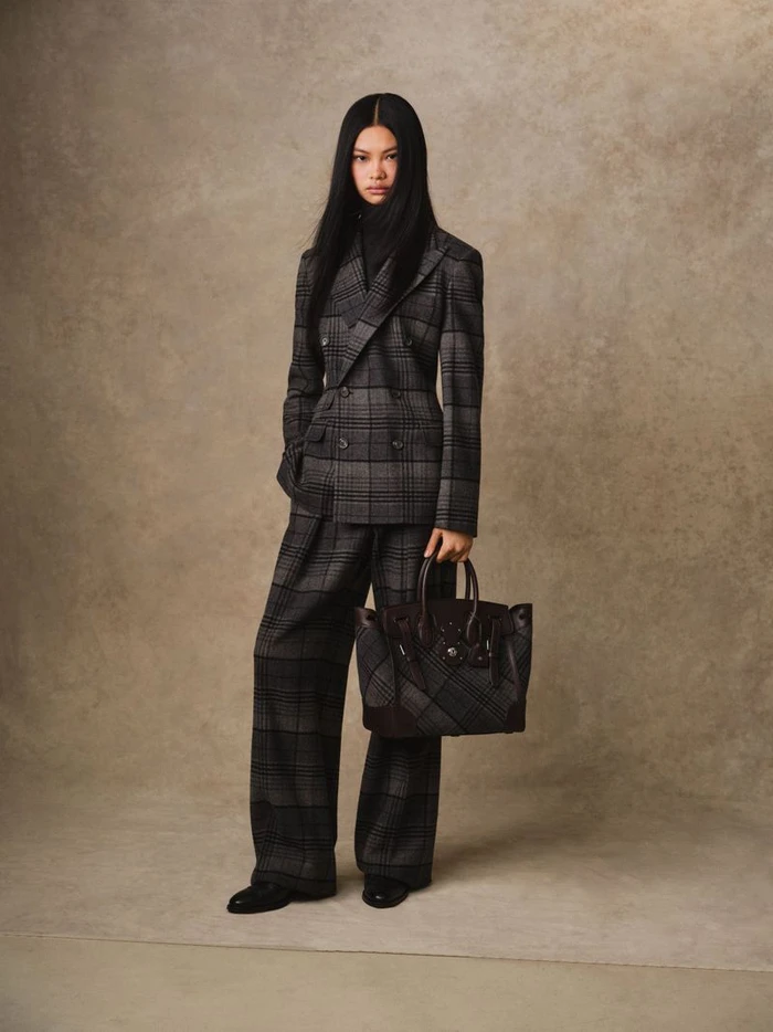 Model Yue Gu standing in a grey plaid double-breasted pantsuit while holding a dark patterned handbag against a textured studio backdrop.