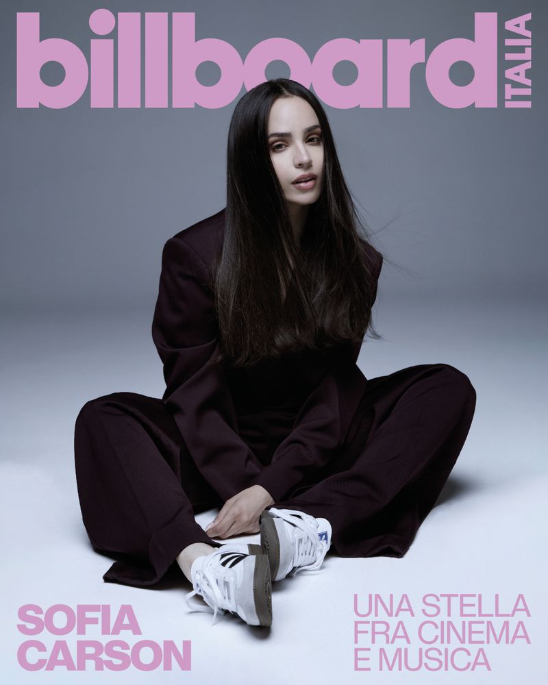 Sofia Carson poses on Billboard Italia cover in a dark suit, seated casually against a light grey background with pink magazine text.