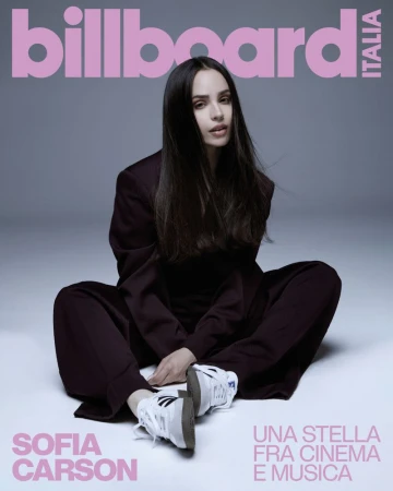 Sofia Carson poses on Billboard Italia cover in a dark suit, seated casually against a light grey background with pink magazine text.