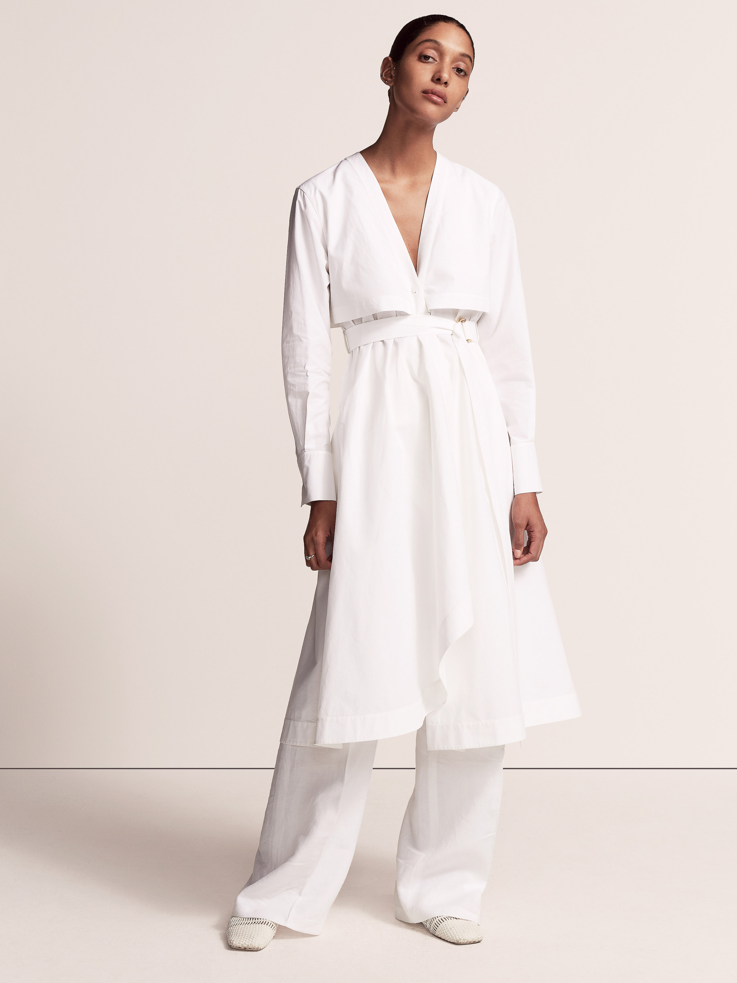 A woman in a white wrap dress and trousers poses in a bright studio environment. The model looks directly at the camera.