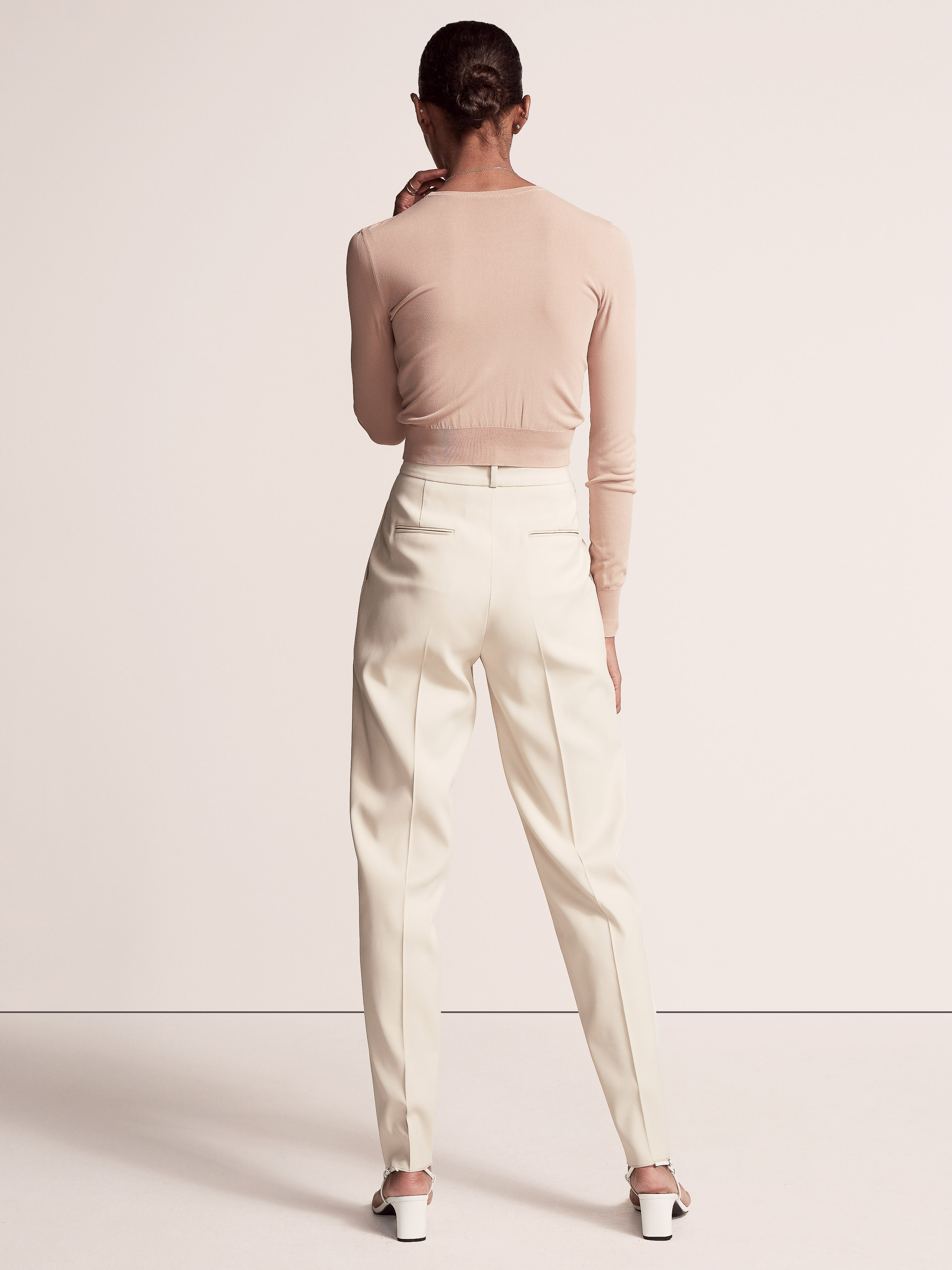 Back view of a woman posing in a long-sleeved top, ivory trousers, and sandals against a neutral studio backdrop. Fashionable and elegant.