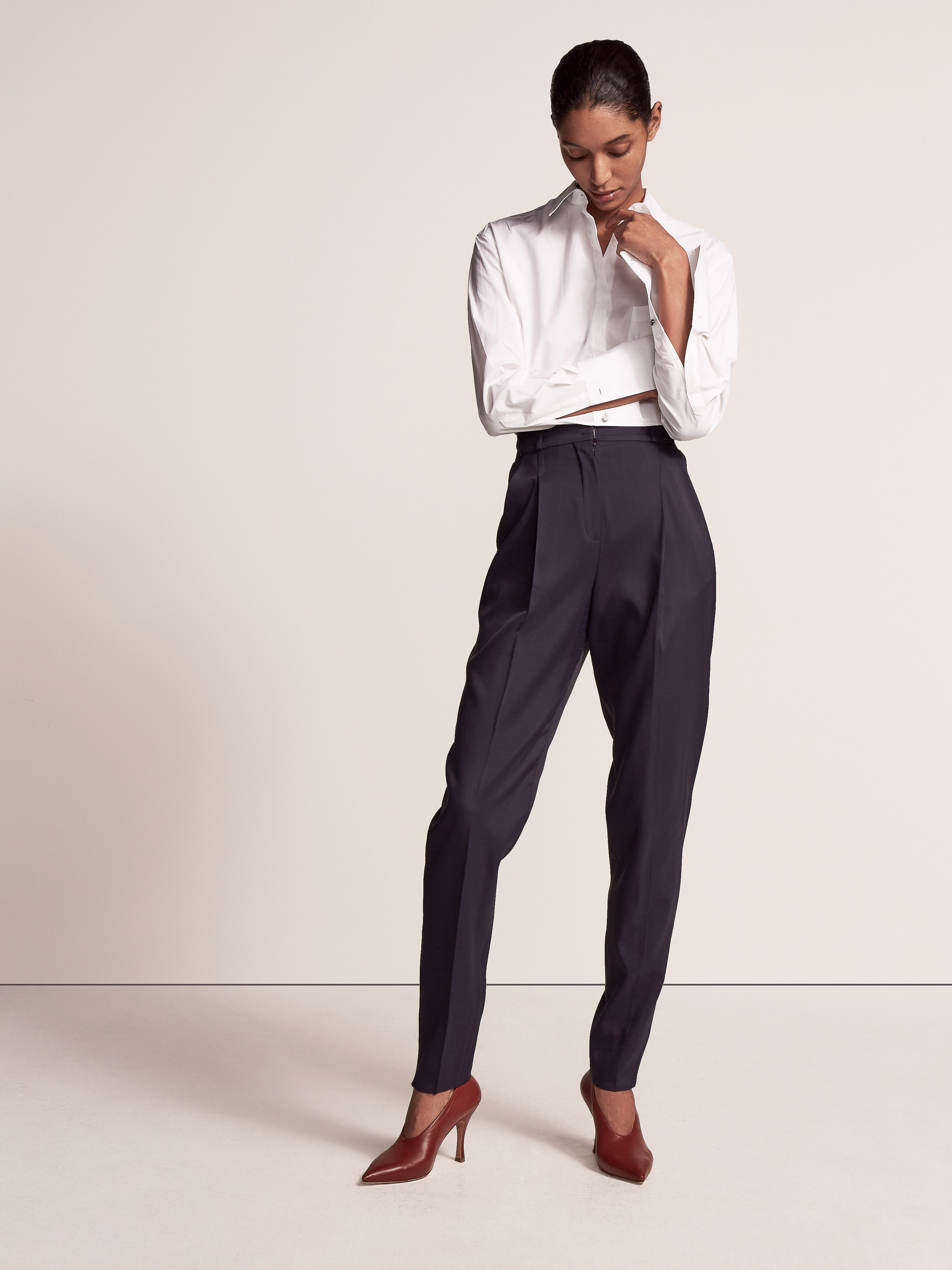 A woman models a long-sleeved white shirt, high-waisted dark trousers, and burgundy heels in a studio shot on a neutral backdrop.