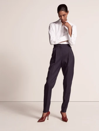 A woman models a long-sleeved white shirt, high-waisted dark trousers, and burgundy heels in a studio shot on a neutral backdrop.