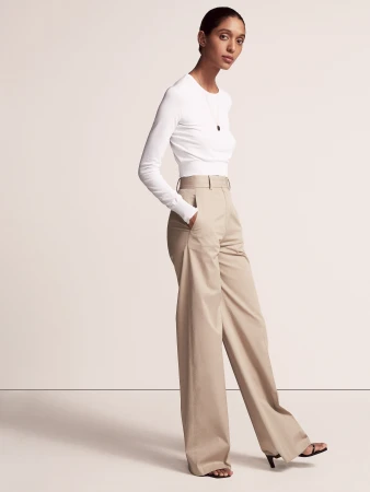 Fashion shot of a slim woman in white long-sleeve shirt and khaki wide-leg trousers standing in a photo studio with bright lighting.