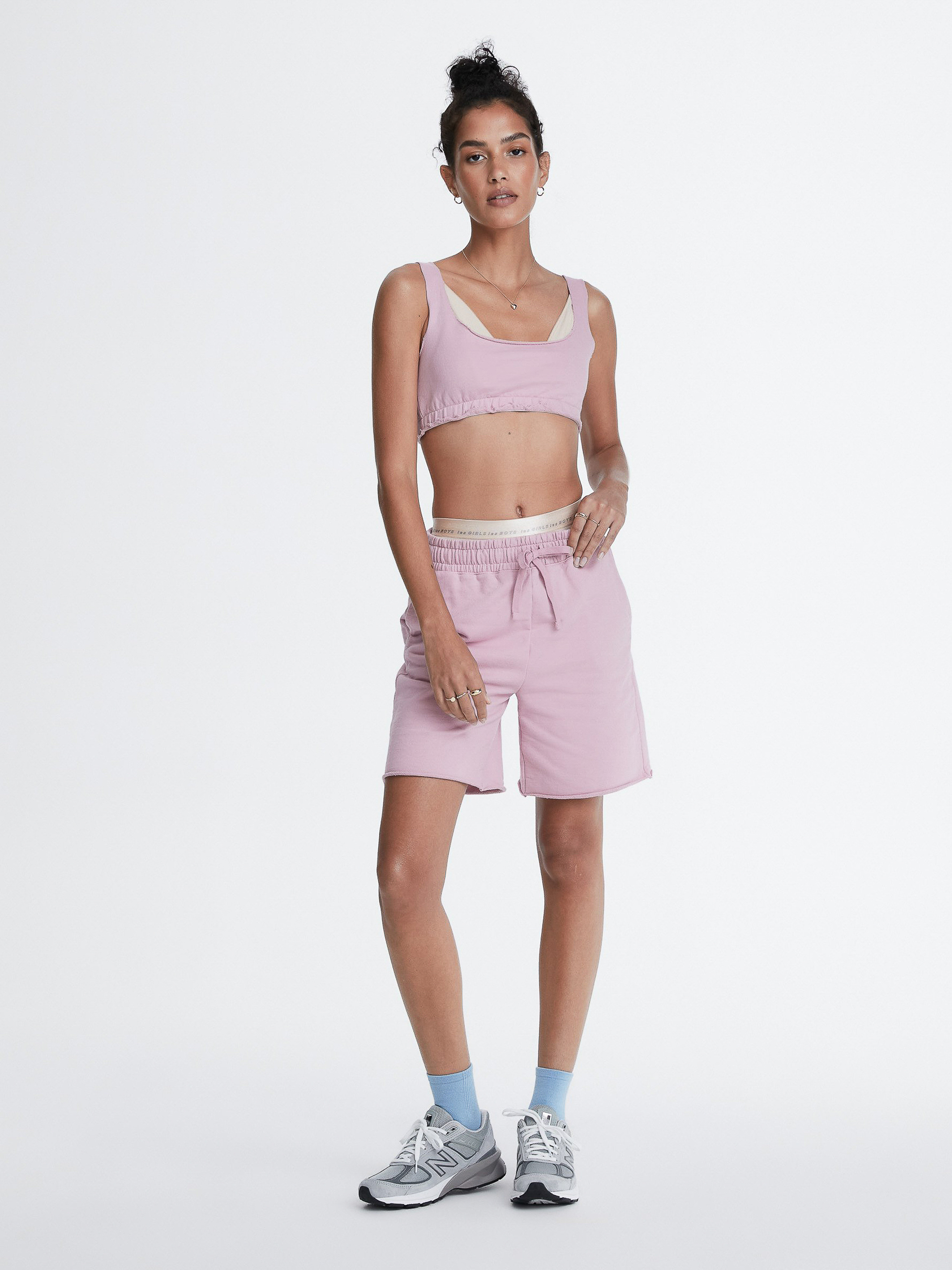 A young adult woman stands in a studio wearing a light pink matching athletic crop top and shorts, paired with running shoes and blue socks.