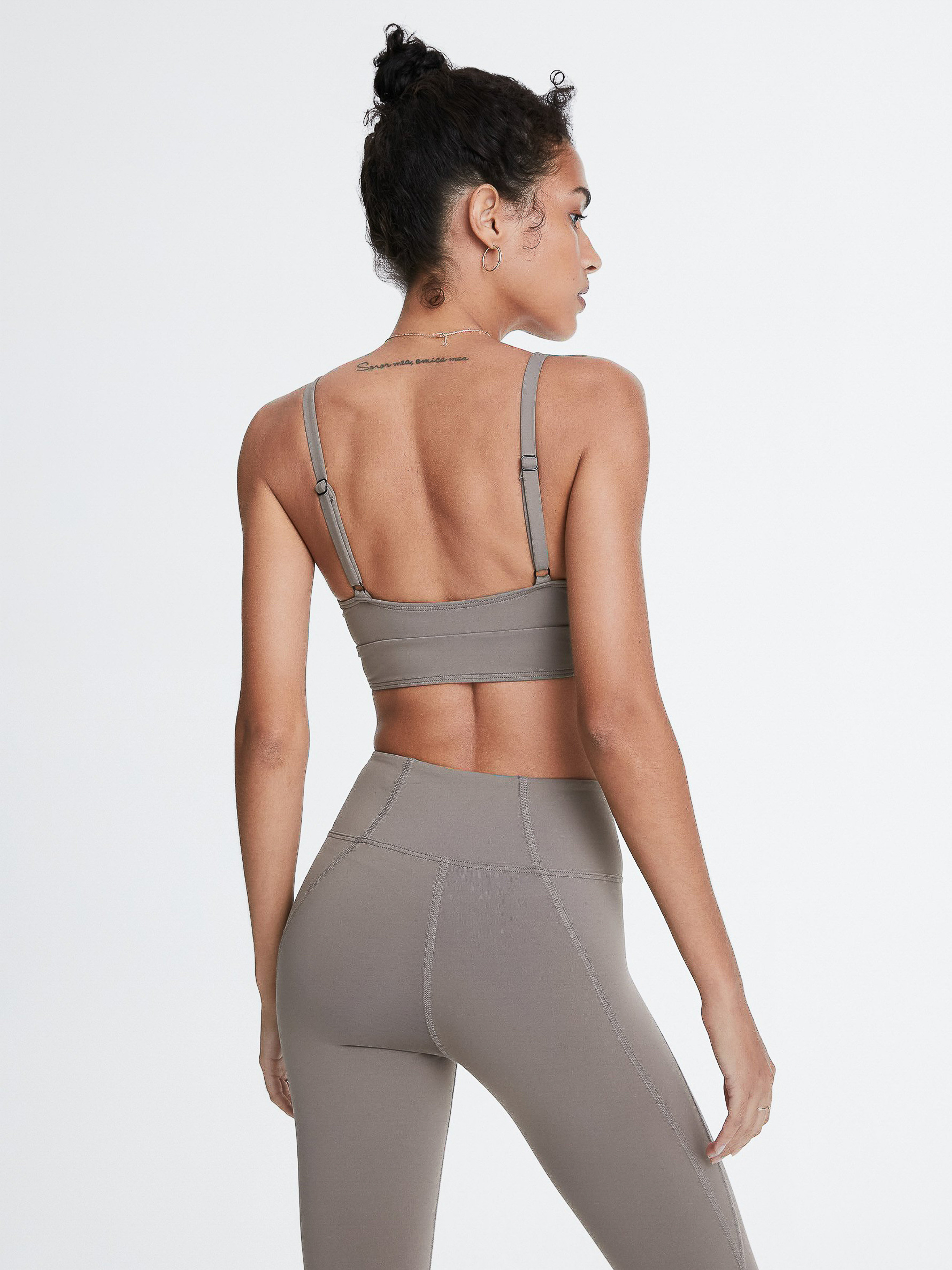 An adult female with brown hair is seen from behind wearing a matching grey sports bra and leggings against a white background.