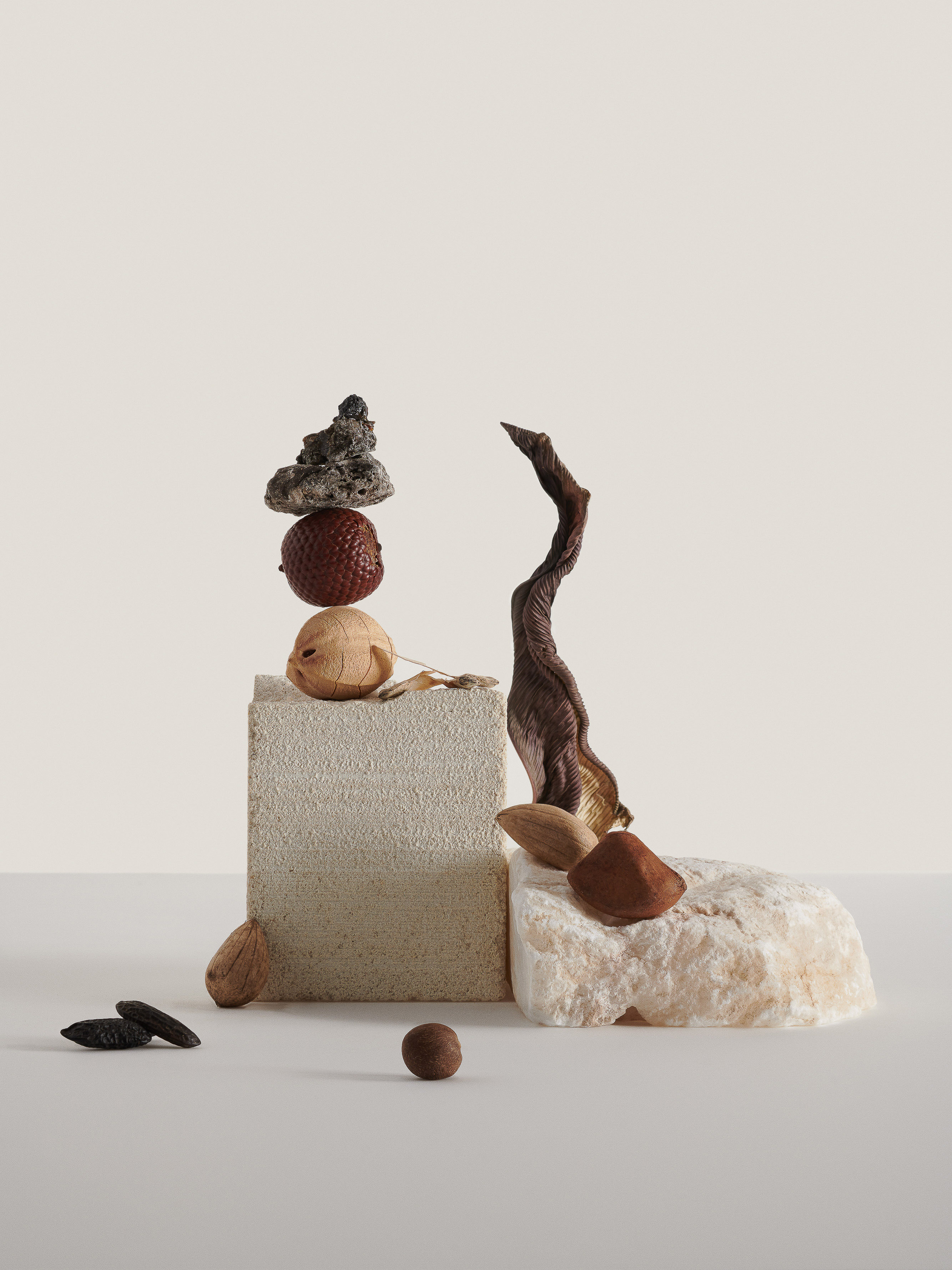 Still life featuring a stacked arrangement of rocks, nuts, and stone fragments against an off-white background. Studio shot.