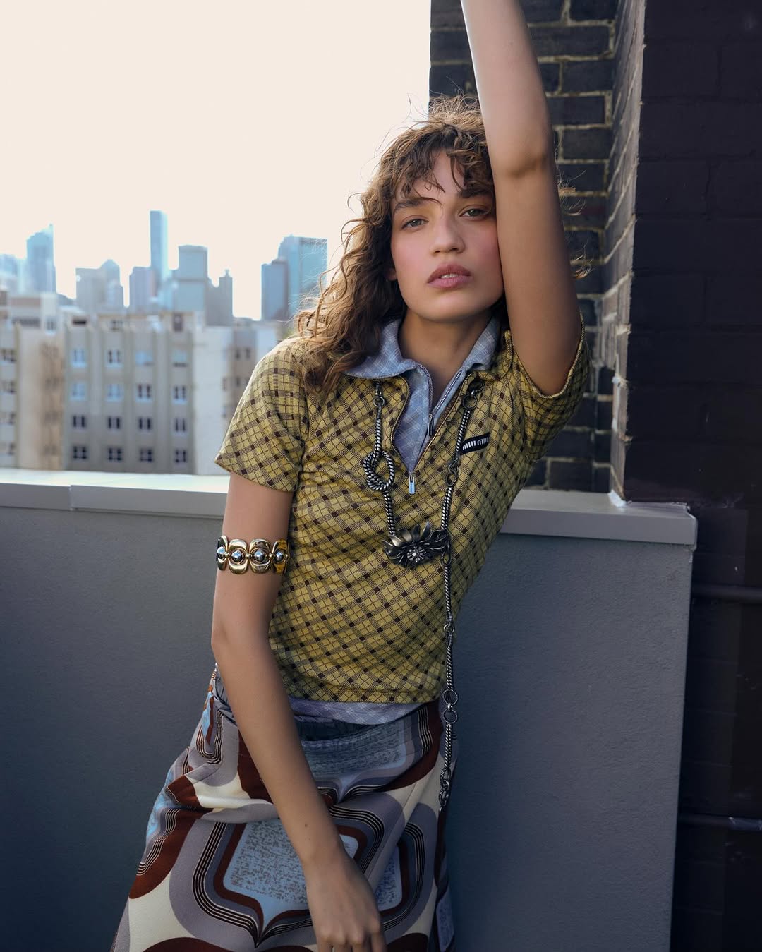 Portrait of a brunette female posing on a rooftop with city skyline. She is wearing a retro shirt and skirt with arm jewelry.