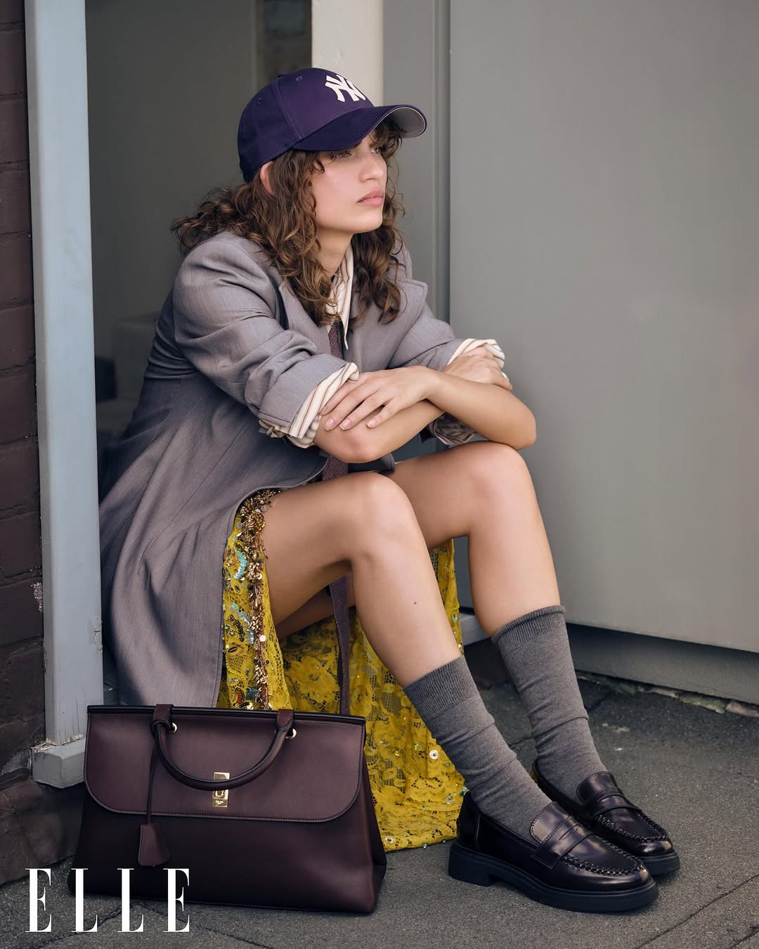 Young woman poses in a grey jacket, baseball cap, and loafers, seated outside a grey door with a burgundy handbag.