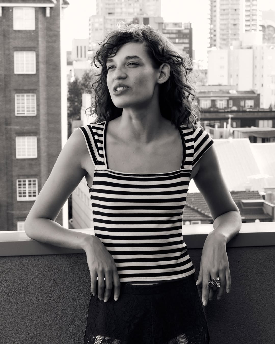 Portrait of a woman with curly hair wearing a striped top, posing on a rooftop with a cityscape in the background, in black and white.